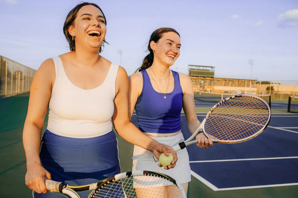 photo of two young women are laughing and holding tennis rackets on a sunny tennis court