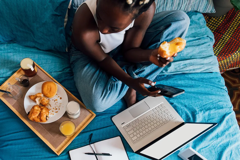 photo of a woman holding mobile phone having breakfast with girlfriend on bed