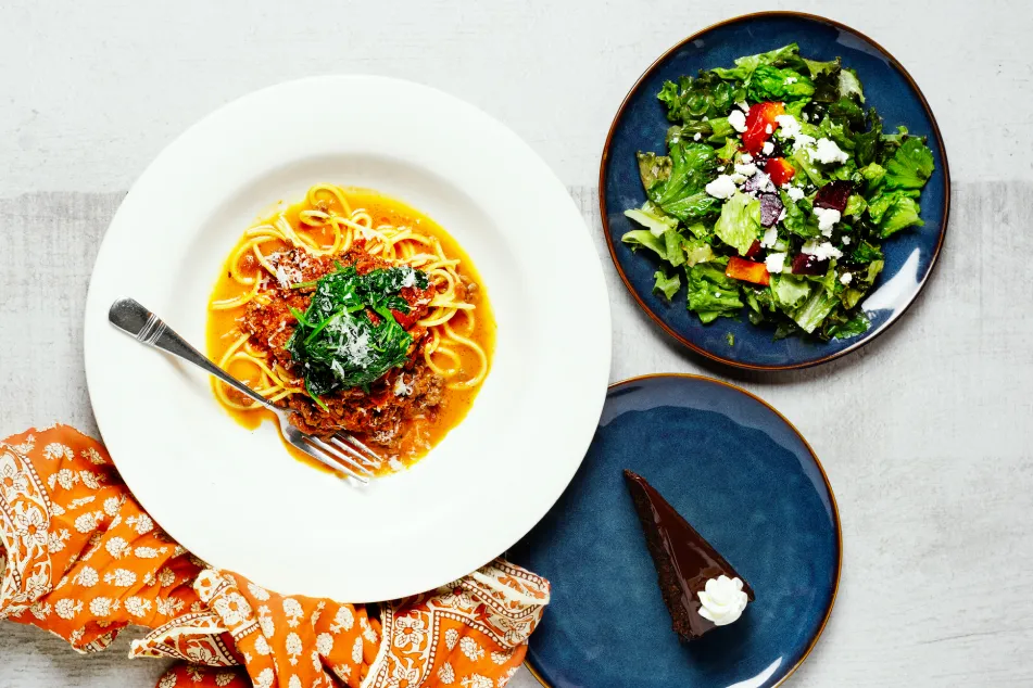 photo of spaghetti bolognese with a plate of salad and a slice of cake